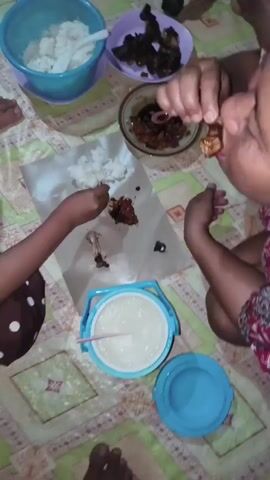 The video shows a group of people eating a meal on the floor. There are several dishes of food, including rice, chicken, and a sauce. The people are using their hands to eat, and they appear to be enjoying their meal. The environment is a home, with a mat on the floor and a chair in the background. The lighting is dim.