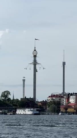 The video shows a view of a waterfront with several tall structures. In the foreground, there is choppy water. In the middle ground, there is a shoreline with buildings and trees. Dominating the scene are three tall towers. The central and largest tower is a swing ride at an amusement park, with several chairs visible hanging from it and slowly rotating. To the right of the central tower is another tall, slender tower with a platform at the top. To the left and further back is a third, even taller and thinner tower. Several boats are visible on the water and docked along the shore. The sky is overcast.