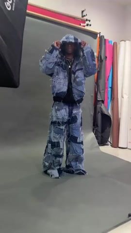 A person stands on a gray floor in front of a gray backdrop wearing a denim patchwork outfit of a hooded jacket and wide-legged pants. The person adjusts the hood of the jacket, then leans forward. To the left is a black softbox light, and to the right are rolls of colored paper backdrops.