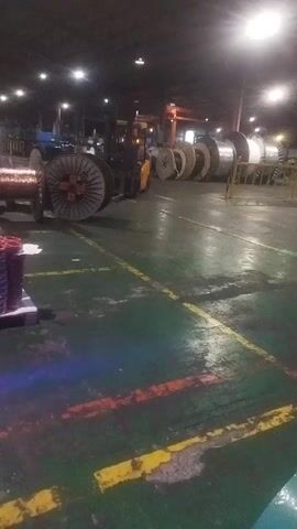 The video captures an indoor industrial setting, likely a warehouse or factory floor, at night or in low light conditions. The floor is marked with green, yellow, and red lines, indicating pathways or designated areas. A forklift is visible in the center of the frame, carrying a large spool of what appears to be copper wire. To the left, another large spool of wire is stationary, with smaller spools stacked on a pallet in the foreground. In the background, a row of numerous large spools, possibly of different materials and sizes, are lined up. Two individuals are seen walking and interacting near the forklift and the large spools. The environment is illuminated by artificial lighting fixtures, and overhead cranes are faintly visible in the upper portion of the frame.
