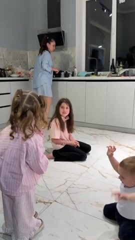 In a brightly lit indoor scene, two young girls and an adult are visible in what appears to be a kitchen. The younger girl, with braided pigtails, wears a pink striped pajama top and faces away from the camera. Another girl, slightly older, sits cross-legged on the tiled floor, dressed in a pink sweatshirt and black pants. She initially smiles but then covers her face with her hand, displaying a strong emotional reaction that suggests embarrassment or laughter. In the background, an adult wearing a light blue pajama set stands near a countertop, partially obscured. The kitchen is equipped with white cabinets and countertops, and various kitchen items are visible in the background. The floor is covered in light-colored tiles with a marble-like pattern. There is music playing.