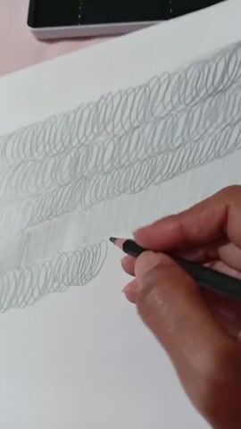 A close-up shot shows a person's right hand holding a black pencil and drawing on a sheet of white paper. The paper is positioned on a pink surface, possibly a table or cloth. The drawing consists of multiple rows of coiled lines, creating a textured effect. In the upper part of the image, a silver metal pencil case is visible, open, revealing several dark pencils inside. To the right of the paper, a few more pencils are scattered, some with visible brand names. The focus is on the hand and the drawing process, capturing the act of creating the coiled patterns. The lighting appears soft and even, illuminating the scene without harsh shadows.