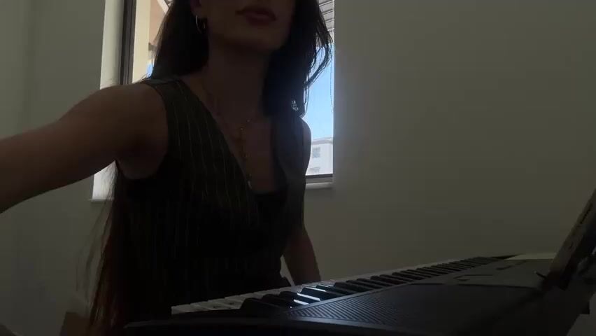 A young woman with long dark hair, wearing a dark vest over a dark top, is seated at a digital piano. She is illuminated by natural light coming from a window to her left. Her hands are positioned over the keys, and she appears to be playing. The woman has jewelry on her left wrist and a ring on her left index finger. The piano has a music stand attached. The background is a plain wall and a window with blinds that are partially open, revealing a glimpse of buildings and a clear sky.