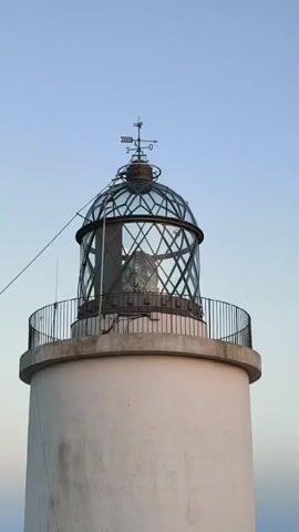 A close-up, vertical shot focuses on the top portion of a white lighthouse under a clear, pale blue sky. The lighthouse has a circular, grey platform surrounding a glass enclosure that houses the lamp. The glass enclosure is framed by crisscrossing metal supports and topped with a decorative weathervane. A black metal railing encircles the platform. A thin black cable extends from the left side of the lighthouse towards the top. The lamp inside the glass enclosure appears to be on, emitting a bright light. A faint, white, circular object, possibly the moon, is visible in the sky to the left of the lighthouse. The lighting suggests it is either dawn or dusk. There are no people visible in the video.
