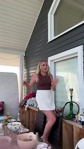 A blonde woman wearing a brown tank top and a white skirt stands on a wooden railing outdoors, holding a shot glass. She is dancing and appears to be at a party. In the background, there is a table with food and drinks, a hookah, and some discarded clothing.
