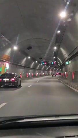 The video shows a view from the driver's seat of a car driving through a tunnel. The tunnel has a concrete ceiling and walls, with rows of lights and ventilation fans visible along the ceiling. Traffic lights displaying green arrows and speed limit signs are mounted on gantries above the lanes. Several cars are visible ahead, including a black sedan in the left lane and other cars further down the road. Red reflective markers are spaced along the sides of the tunnel. The road surface is asphalt, and lane markings are white. The time stamp in the video indicates it is 00:00 to 00:05.