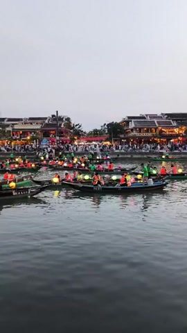 The video shows a river scene at dusk, possibly in Vietnam, with several small boats carrying passengers. The boats are adorned with lit lanterns, creating a festive atmosphere. People in the boats are wearing orange life vests. The river is surrounded by buildings and trees. There are many people walking along the riverbank. The sky is overcast, contributing to a soft light over the scene.