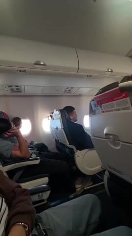 The video shows the interior of an airplane cabin. Passengers are seated in rows of seats. Some passengers are visible in the foreground and mid-ground, facing away from the camera or looking to the side. Overhead compartments are open in some places. Magazines are visible in seat pockets. The lighting appears to be artificial, with some bright lights visible on the ceiling. The windows show a view of the sky or clouds.