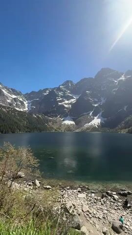 The video shows a scenic view of a lake surrounded by mountains on a sunny day. The lake is dark green with clear water near the rocky shoreline. The mountains are tall and rugged, with patches of snow visible on their slopes. Evergreen trees cover parts of the lower mountain slopes. The sky is a clear blue with the sun shining brightly, creating a lens flare effect.  Some people are visible near the shoreline. The camera is relatively still, providing a stable view of the landscape.