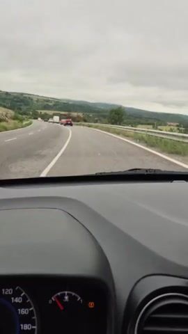 The video shows the interior of a car driving on a road. The camera is positioned behind the dashboard, looking forward through the windshield. The road is paved and has white lane markings. On the left side of the road, there are rolling hills covered in green vegetation. On the right side, there is a metal guardrail and some trees, some of which appear to be burnt. The sky is overcast with gray clouds. There are a few other vehicles visible on the road in the distance, including cars and a truck. A phone mount is visible on the dashboard in the foreground. The speedometer is partially visible at the bottom left of the screen, showing the needle pointing to a speed below 100. A dashboard warning light, possibly for the engine or emissions system, is illuminated. The video captures the motion of the car as it travels along the road, with the landscape passing by.