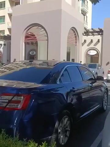 The video begins with a shot of the rear of a dark blue sedan parked in a parking lot. The building behind the car has a beige facade with arched entrances and decorative awnings. To the right, a white car is visible near an entrance. The camera pans slightly to the left, showing more of the building and the sky. Further to the left, a tall, modern building with multiple balconies is visible. In the foreground, a dense green hedge lines the edge of the parking lot. The camera continues to pan, revealing a green SUV parked next to a silver car in front of the building. The scene appears to be a well-maintained outdoor area with a mix of modern and traditional architectural styles.