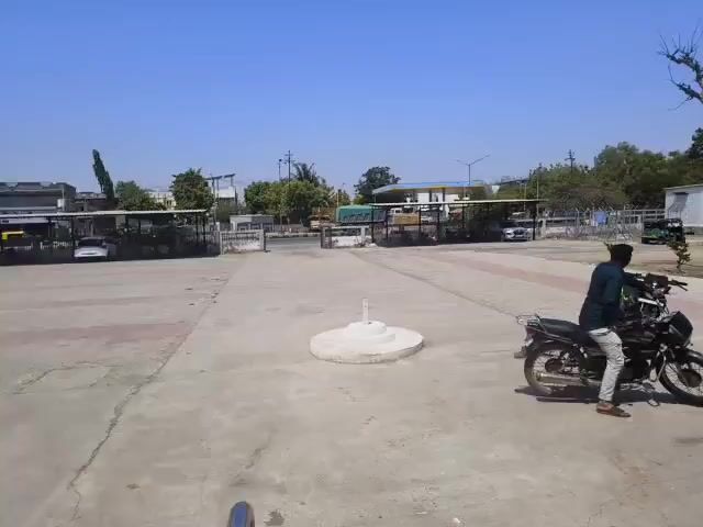 The video shows a parking lot on a sunny day. Two young men are present, one getting on a motorcycle. A second man is walking with a backpack. A yellow and green auto rickshaw and other parked motorcycles are visible in the lot. A white building is on the right side of the frame. A tree without leaves stands nearby. In the background, a fenced area is filled with parked vehicles.