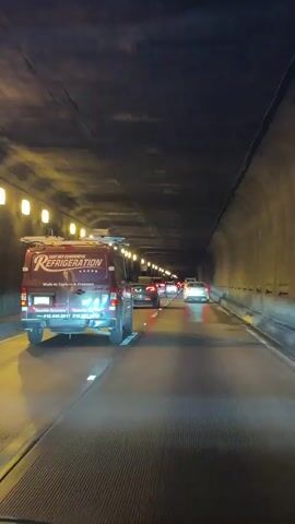 A car POV shot shows traffic moving slowly through a tunnel. The tunnel is illuminated by overhead lights and the taillights of the cars. The road surface is wet. The video begins with a commercial refrigeration van in the foreground, followed by several cars, including a Tesla and a Lexus. The camera is moving forward, and the traffic appears to be congested. The footage is grainy and appears to have been recorded at night or in low light conditions.