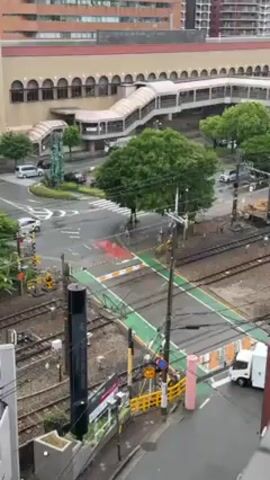 Here is a description of the video:

The video shows a train crossing a road in Japan. The train is blue and silver. It is travelling along a set of tracks that crosses the road. There is a green crosswalk marking for pedestrians to use. The road is wet from a recent rain. The video is taken from an elevated position, looking down at the scene. A large building is visible in the background, which seems to be a railway station, based on the overhead pedestrian bridge. There are cars and a small white truck driving on the road.