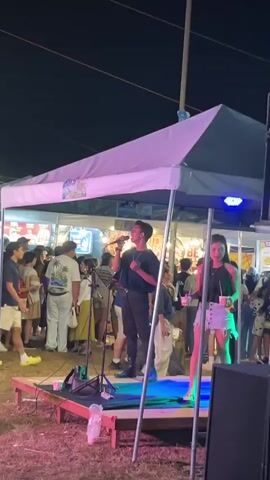 A young man sings into a microphone on a small stage under a large canopy. He is wearing a black t-shirt and black pants. A young woman stands next to him, wearing a black tank top and denim shorts. Behind them, a crowd of people are visible, many standing and watching the performance. In the background, there are stalls and lights, suggesting an outdoor market or festival at night. To the right of the stage, large speakers are stacked. The ground is covered in dry grass or dirt.
