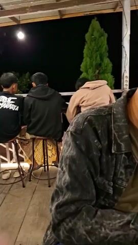 The video opens with a close-up of a young man wearing a denim jacket and a green t-shirt. He has short, styled hair and a mustache. The camera then pans to the right, showing a drink with ice and a straw on a table. The background reveals a nighttime outdoor setting with numerous string lights illuminating a multi-level building. People are seated at tables on the ground floor and on the upper levels of the building, and some are gathered in an outdoor seating area with white benches and picnic tables. Several motorcycles are parked in the background. The scene appears to be a busy cafe or restaurant.