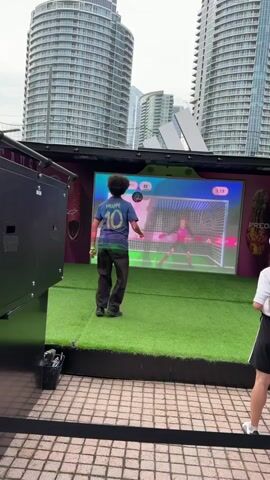 A person with dark curly hair wearing a blue soccer jersey with the number 10 and the name Mbappe on the back is playing a soccer video game. The person is standing on a green turf mat in front of a large screen displaying a soccer game. The screen shows a virtual soccer goal with a goalie in a pink uniform. The player kicks a virtual ball towards the goal. The environment appears to be outdoors with tall buildings in the background. Another person is standing to the right, partially visible and wearing a white shirt and black shorts. There are also some bubbles floating in the air.