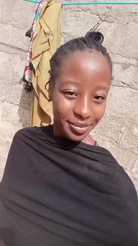 A person with dark skin, wearing a black shawl, stands in front of a textured wall. The person's hair is pulled back in a bun. A line of clothes hangs on the wall to the person's left. The person smiles and blows a kiss towards the camera. The scene is shot in natural light.