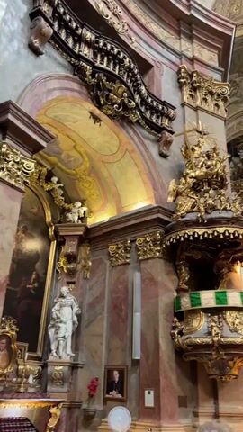 The video pans across the interior of a grand, ornate church. The architecture is a Baroque style, with abundant gold leaf, marble, and intricate carvings. A prominent feature is a richly decorated pulpit on the right side of the frame, adorned with gilded figures and a green and white fabric banner. To the left, a statue of a robed figure stands on a pedestal. Further in the background, an altar is visible with a painting above it. The ceiling is high and vaulted, with painted frescoes and decorative moldings. The lighting is somewhat dim, typical of a church interior, with some areas illuminated by natural light from arched windows and some by artificial lights. Several people are present, mostly seen from the back, standing and observing the church's interior. They appear to be tourists or visitors. The atmosphere is solemn and reverent, a classic depiction of the interior of a historic European church.