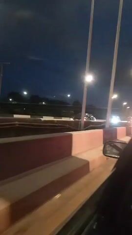 The video shows a dark, possibly nighttime or dusk, scene with streetlights illuminating the area. A car is moving along a road, and the camera is positioned inside the car, looking out. In the distance, there are several cars, some with their headlights on. There appears to be a person in an orange shirt walking on a raised area or walkway to the left of the road. The road is bordered by concrete barriers with red and cream colored sections. Power lines and poles are visible in the background. The overall impression is of a busy road or highway at twilight or night.