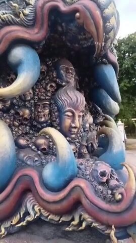 The video displays a close-up view of a detailed, colorful sculpture. The sculpture is dark gray and features numerous sculpted human faces and skulls embedded in its surface. Vibrant red and blue wavy forms are prominent, some of which appear to be stylized tentacles or flowing fabric. These elements are intertwined with the faces and skulls, creating a complex and somewhat macabre aesthetic. The sculpture is outdoors, with hints of greenery and a cloudy sky visible in the background, suggesting it is part of a larger monument or art installation. The camera pans across the sculpture, focusing on the intricate details of the sculpted faces and the dynamic curves of the colored elements.