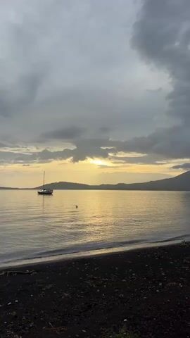 A solitary sailboat is anchored in calm waters near a dark sand beach under an overcast sky. The sun is partially visible, casting a golden glow across the water and illuminating the distant, tree-covered hills. The waves are gentle, lapping at the shore.