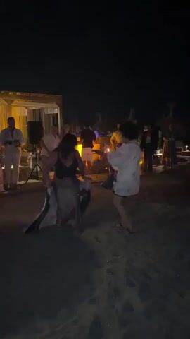This media file captures a lively nighttime scene on a sandy beach, likely at Playa Del Sol-Villacana, Spain. The primary focus is on two individuals dancing energetically in the foreground. One person, wearing a dark top and a light-colored skirt, is in a low, dynamic pose, holding a green and white striped cloth. Facing them, another person in a light-colored, patterned shirt is also in motion, seemingly engaged in a dance with their companion.

In the background, a group of people are gathered, suggesting a party or event. There are several straw-thatched structures and a wooden building with open sides, illuminated by warm lights, hinting at a resort or beach club setting. A DJ booth with equipment and a glowing fire pit or illuminated centerpiece add to the festive ambiance. The people in the background appear to be socializing and enjoying the music.

The time of day is clearly night, indicated by the dark sky and artificial lighting. The sandy ground suggests a beach location, and the presence of palm-like structures reinforces a warm-weather, potentially Mediterranean, setting. The mood appears to be joyful and celebratory, with the dancers exuding enthusiasm. No visible text is present in the media file.