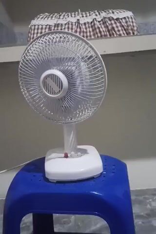 A white desk fan is sitting on a blue plastic stool. The fan is on and its blades are spinning. The background is a wall with a shelf above the fan. There is a checkered fabric draped over the shelf. The lighting is dim and appears to be artificial.