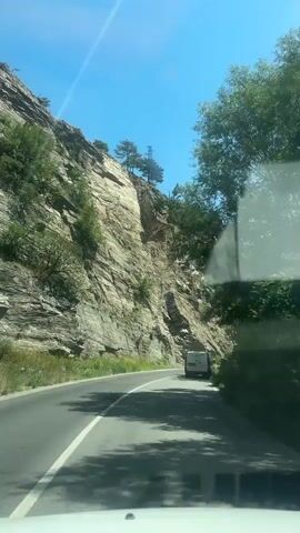This video shows a view from a vehicle driving along a paved road that curves to the right. On the left, a steep, rocky cliff face rises dramatically, covered in sparse vegetation and trees near its summit. The cliff is layered and appears to be made of light-colored stone. On the right side of the road, trees and dense green foliage line the route, creating a shaded canopy. A white van is visible driving ahead on the road. The sky is clear and bright blue, with a few wispy white clouds. The sun is shining, indicated by the bright light and the shadows cast on the road. The time of day appears to be midday, given the strong sunlight. The environment suggests a mountainous or canyon-like setting, typical of a scenic route in a rural or natural area. The overall mood is one of travel and exploration through a picturesque landscape. No visible text is present in the video. The location is identified as Vetren Dol, Bulgaria.