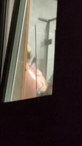 A person with blonde hair pulled back in a bun is showering. They are facing away from the camera, and their back is visible. The person is standing in a shower stall with glass doors. There is a showerhead and faucet visible on the wall. The lighting is dim, and the image is somewhat blurry.