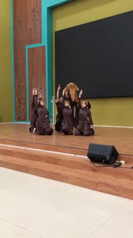 Six young people in brown outfits with head coverings are performing a dance on a wooden stage. They are in various kneeling and crouching positions, with their arms raised. One person in the center, slightly taller, is holding a large, light-colored stuffed animal above their head. Another person in the back is holding the stuffed animal, while the rest of the group is performing synchronized movements. The stage has a wooden floor and is elevated by wooden steps. A black speaker is visible on the lower steps. The background features a large dark screen and a wall with wooden paneling and teal accents.