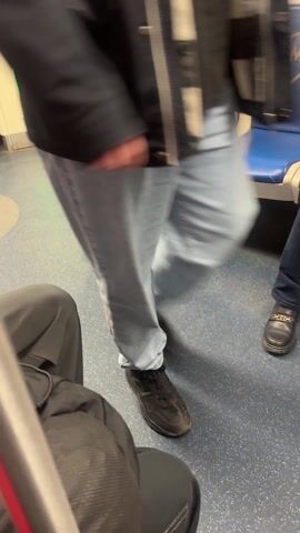The video shows the interior of a subway car. A person wearing a white puffy jacket, gray sweatpants, and gray sneakers is sitting on a blue and white seat, holding a white bag and looking at their phone. The subway car floor is blue with silver speckles. Another person wearing black pants is sitting in the foreground, partially out of view.  A small portion of another person's leg wearing a black shoe is also visible. The camera is at approximately waist height.