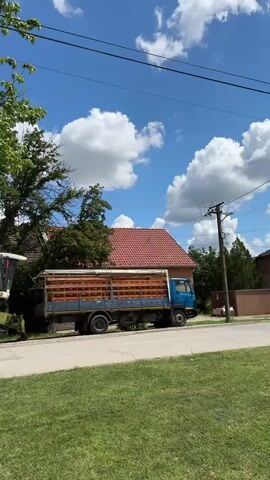 The camera pans left across a rural or suburban street scene on a bright, sunny day. A large green and white combine harvester, labeled "Claas Mega 204," is parked on the left side of the road. To its right, a light blue flatbed truck with wooden side rails is also parked. A large, leafy green tree partially obstructs the view of the vehicles as the camera pans. In the background, a house with a red-tiled roof is visible, along with power lines and a utility pole. Further down the street, a white van is briefly seen. The foreground consists of a green grass verge and a paved road.