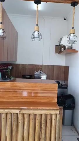 The video shows an indoor scene of what appears to be a kitchen or food service area. The camera is positioned at a medium distance, capturing a counter made of wood with a glossy finish in the foreground. The base of the counter is constructed from vertical bamboo poles. Behind the counter, a backsplash made of wood paneling is visible, with various items placed on it, including what looks like cooking utensils and appliances. To the right of the counter, there is a stove and a trash can. Two pendant lights with a rustic design, featuring cages and rope cords, hang from the ceiling. The walls are painted white, and a shelf is mounted on the back wall, holding some containers. There is also a security camera visible on the ceiling. The lighting in the room is bright, likely from the overhead lights and possibly natural light from an unseen source. No people are visible in the video, and no specific actions or interactions are depicted. The overall impression is of a clean and functional space with a natural, possibly tropical, aesthetic due to the use of bamboo.