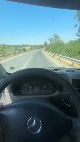 The video shows a first-person view from inside a vehicle, focusing on the dashboard and the road ahead. The driver's hands are visible on the steering wheel. The vehicle appears to be a Mercedes-Benz van, indicated by the logo on the steering wheel. The dashboard displays the speedometer, tachometer, and various warning lights. The speedometer needle is around 90 km/h, and the tachometer needle is around 2500 rpm. There are three orange warning lights illuminated: a brake warning light, an ABS warning light, and an ESP warning light. There is also a red warning light, possibly an engine warning light. The digital display shows an odometer reading of 299096 km and a trip meter reading of 640.3 km. The vehicle is driving on a paved road with two lanes in each direction, separated by a white line. There is a guardrail on the right side of the road, and trees and vegetation line the roadside. In the distance, there are houses and more vegetation. The sky is clear and blue, indicating a sunny day. A speed limit sign of 60 is visible on the right side of the road. The video captures the motion of the vehicle as it travels along the road.