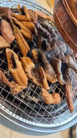 A close-up, overhead shot shows an assortment of meats and what appears to be eel grilling on a circular metal grate over hot coals. The food is sizzling and some pieces are visibly charred. Smoke rises from the grill, obscuring some of the food in the background. The background is blurred and out of focus, suggesting the shot is taken at a restaurant or a barbecue. There are no people visible in the frame.