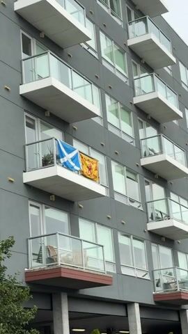 The video shows the side of a modern apartment building with multiple balconies. Two flags are hanging from the railing of one of the balconies. The flag on the left is the Scottish flag, a white saltire on a blue background. The flag on the right is yellow with a red lion rampant.