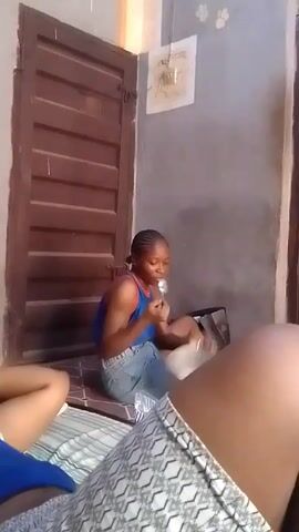 Here's a description of the video:

The video shows a sparsely furnished room. A young woman with braided hair sits on a mattress, wearing a blue top and denim shorts. She's looking towards the right of the frame and seems to be talking or eating. There are a few items scattered around her on the mattress. Another person is lying down in the foreground, only their torso and legs visible. They are wearing a blue shirt and patterned shorts. A closed wooden door is visible in the background to the left. The walls are painted a light gray, and there are some electrical fixtures and smudges visible on the wall behind the seated woman. The camera moves around a bit, and some furniture is seen in the background.