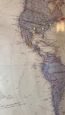 The video shows a world map mounted on a wall, likely made of wood judging by the texture visible at the top of the frame. The map is covered with numerous pushpins of various colors, including white, yellow, and red. These pins are used to mark locations on the map. The narrator states in Russian that these are places where her husband has traveled. The camera pans across the map, showing pins in North America, Central America, South America, Africa, and parts of Europe and Asia, suggesting extensive travel. The lighting in the room appears to be artificial, casting a warm glow on the map and creating some reflections on its surface.