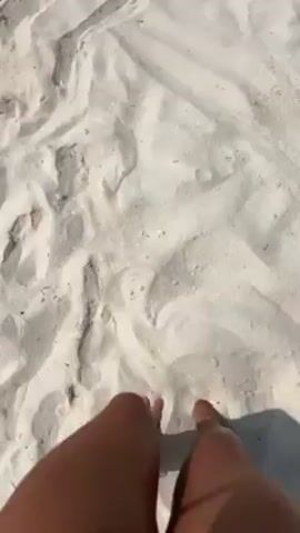 The video shows a beach scene from a first-person perspective, with the camera focused on the person's feet initially covered in sand.  The camera then pans up to reveal the turquoise ocean and white sand beach.  People are swimming in the water and enjoying the beach.  The sky is overcast, but the overall scene is bright.  The camera pans from right to left along the beach. The person has an ankle bracelet on their left ankle.