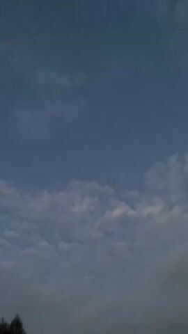 The video shows a wide shot of a cloudy sky at dusk. There are a few scattered clouds, some of which are tinged with pink. A power line stretches diagonally across the frame from left to right. At the bottom of the frame, the tops of trees are visible. The camera appears to be moving slightly, causing some blur in the image, especially in the lower portion. The overall mood is serene and peaceful, with the colors suggesting the end of the day.