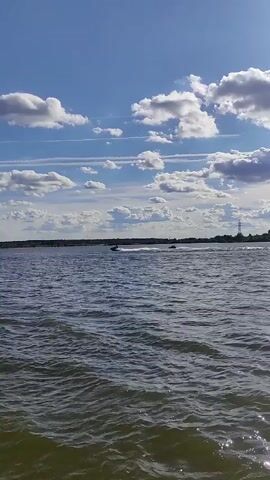 The video shows a wide shot of a body of water on a sunny day. The sky is blue with fluffy white clouds. On the water, two jet skis are visible in the distance. The jet ski on the left is kicking up a spray of water as it moves at a high speed. The jet ski on the right appears to be moving slower and is further away. The water is choppy, indicating a light breeze. The background shows a long stretch of land with trees and a tall pylon, suggesting a rural or semi-rural setting. The overall mood is one of leisure and recreation on a pleasant day. The timestamp indicates the video was recorded between 00:00 and 00:02.