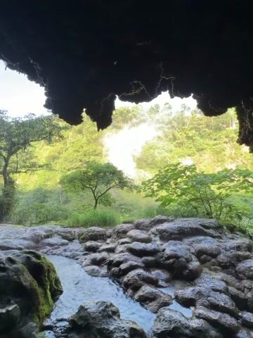 The video pans across a natural scene viewed from inside a dark cave or overhang. In the foreground, there are wet, rocky surfaces with moss. A small stream of water flows through the rocks. Beyond the rocks, a lush green landscape is visible, with trees and foliage. In the distance, there is a waterfall creating a misty spray that catches the sunlight, creating a bright, ethereal glow. The sky is visible above the mist. The overall impression is of a hidden, serene natural environment.