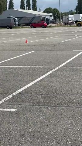 The video shows a motorcyclist performing a maneuver on a paved surface, likely a training ground or parking lot. The motorcyclist, wearing a helmet and protective gear, is on a dark-colored motorcycle with red accents. They are navigating around orange traffic cones. In the background, there are buildings, parked vehicles including a van and cars, and trees under a cloudy sky. The motorcyclist appears to be practicing precise control, possibly a wheelie or a tight turn.