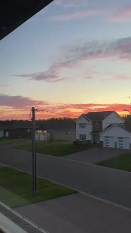 This is a video clip of a suburban street at sunset in Dieppe, Canada. The sky is a vibrant mix of pinks, oranges, and purples, with scattered clouds. Several houses are visible along the street, varying in architectural style, from traditional two-story homes to more modern designs. Some houses have attached garages, and a few cars are parked in driveways. A power line with a utility pole runs along the street, partially obscuring the view. The overall scene conveys a sense of peacefulness and the beauty of a summer evening. The time of day is clearly sunset, indicated by the colorful sky. There is no visible text in the video.