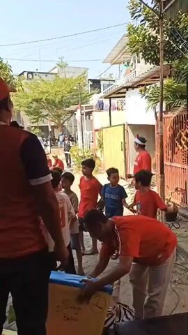 A group of children in red shirts and some adults are gathered outdoors. In the foreground, a man in a red shirt and hat is holding a yellow box with the number 2 and some writing on it. Several children are standing around him, and some are looking at the box. In the background, more people are sitting and standing, and there are buildings and trees. A red and white Indonesian flag is visible. It appears to be a community event or celebration.