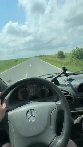 The image captures a first-person perspective from the driver's seat of a vehicle, looking straight ahead through the windshield. A person's right hand is visible, resting on a gray Mercedes-Benz steering wheel, which prominently displays the brand's logo. The dashboard and instrument cluster, including a speedometer (showing markings up to 200, likely km/h), are in view.

The vehicle is driving on a straight, paved two-lane road that stretches into the horizon. White dashed lines mark the center of the road. On either side, vast green fields extend under a bright blue sky dotted with white, fluffy clouds, indicating clear and sunny weather. A distant car can be seen approaching in the far distance on the same road. A phone mount or similar device is attached to the dashboard on the passenger side. The scene depicts a calm drive through a rural landscape, consistent with the context of Madara, Bulgaria.