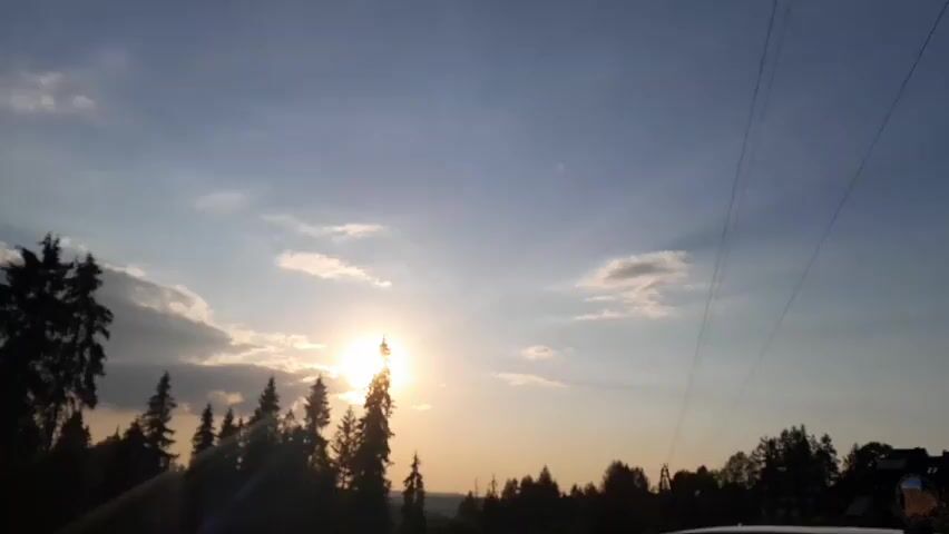 The video captures a sunset over a forested area, likely a rural landscape. The sun is low in the sky, casting a warm, golden light that filters through scattered clouds and illuminates the tops of tall evergreen trees. The trees are silhouetted against the sky, creating a dramatic contrast. In the distance, a faint outline of buildings or structures can be seen on a rise. The sky transitions from a bright blue at the top to a softer yellow and orange near the horizon, a typical display of sunset colors. Several thin, dark lines stretch across the upper portion of the frame, suggesting power lines or telephone wires. The overall atmosphere is serene and natural, evoking a sense of peace and the end of the day.