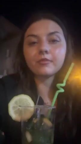 A close-up, slightly blurry video shows a young woman with dark hair and dark clothing drinking from a tall glass filled with a green beverage, ice, mint leaves, and lime slices. The glass has a salt rim and is garnished with a slice of lemon. The woman is holding a green straw and appears to be sipping the drink. The background is dark with a hint of building lights. The overall lighting is dim.
