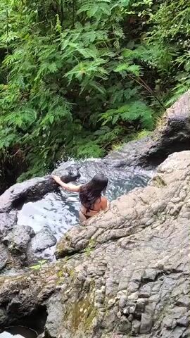 The video shows an outdoor scene with dense green foliage surrounding a rocky area with small pools of water. A woman with long dark hair wearing a dark bikini is in one of the pools. The camera pans to the right to reveal three more women standing on the rocks next to the pool. One woman has red hair and wears a black bathing suit, another wears a white bikini, and the third has long dark hair and wears a yellow swimsuit. One of the women holds a camera.